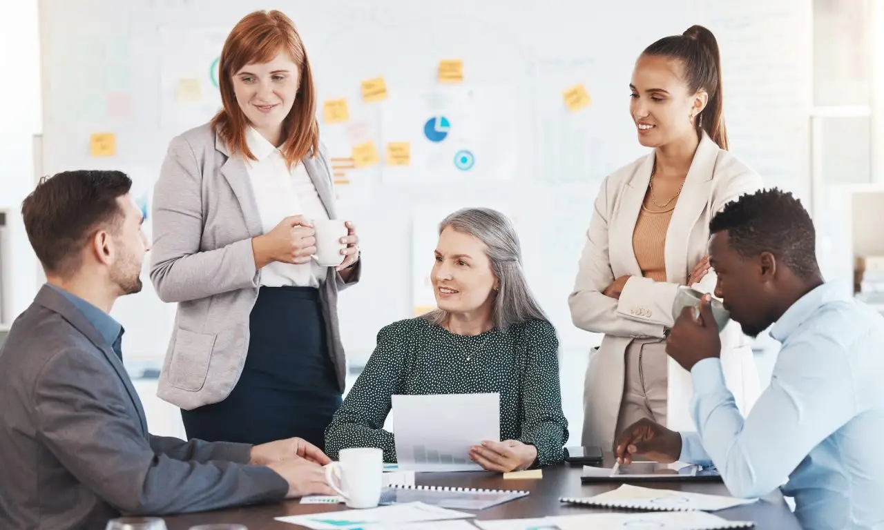 Five business professionals—three women and two men—are having a discussion around a table in an office meeting room. Papers, coffee cups, and documents are on the table, and a whiteboard is in the background. STG-Health