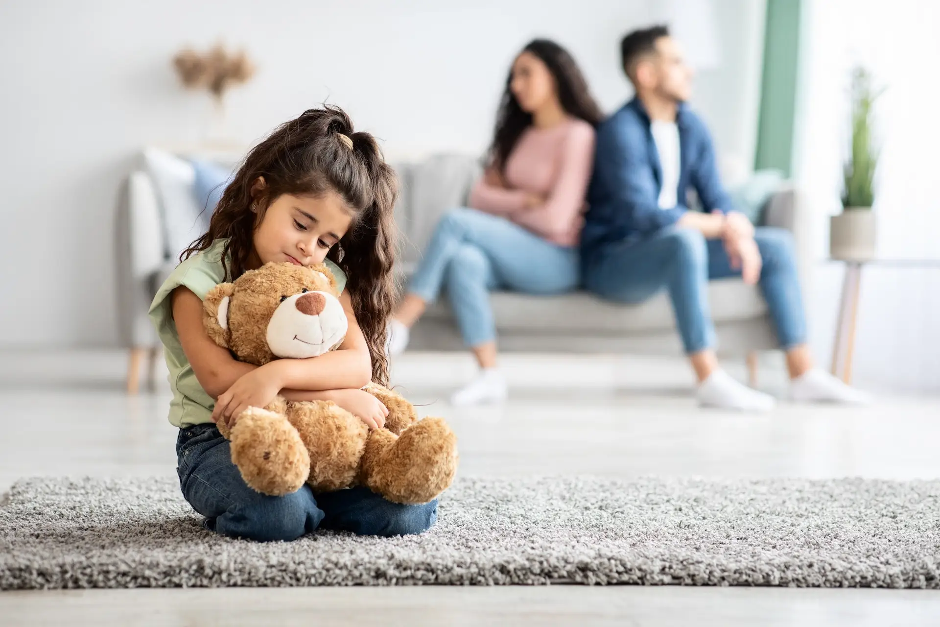 A young girl sits on a rug, hugging a teddy bear and looking sad. In the background, a man and woman sit on a sofa, turned away from each other, appearing upset. STG-Health