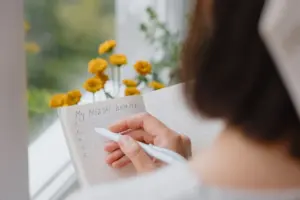 A person writes My MENTAL health at the top of a grid notebook, holding a pen and sitting by a window with yellow flowers visible outside. The page has a numbered list from one to six. STG-Health