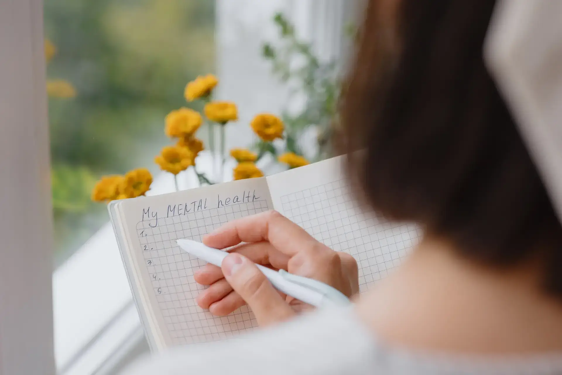A person writes My MENTAL health at the top of a grid notebook, holding a pen and sitting by a window with yellow flowers visible outside. The page has a numbered list from one to six. STG-Health