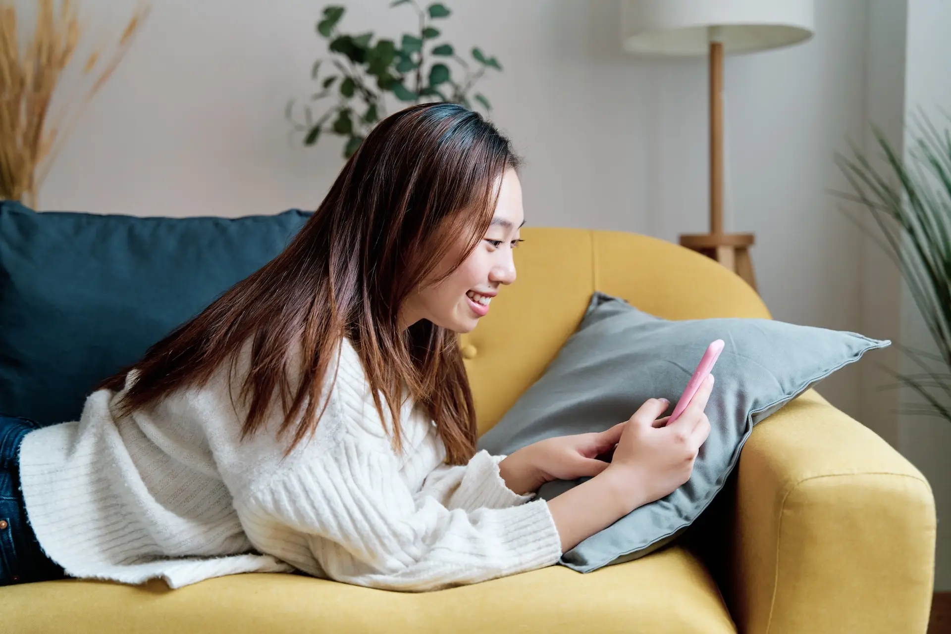 A young woman lies on a yellow sofa, smiling while using a pink smartphone. She wears a white sweater and has long brown hair. There are cushions, plants, and a lamp in the background. STG-Health