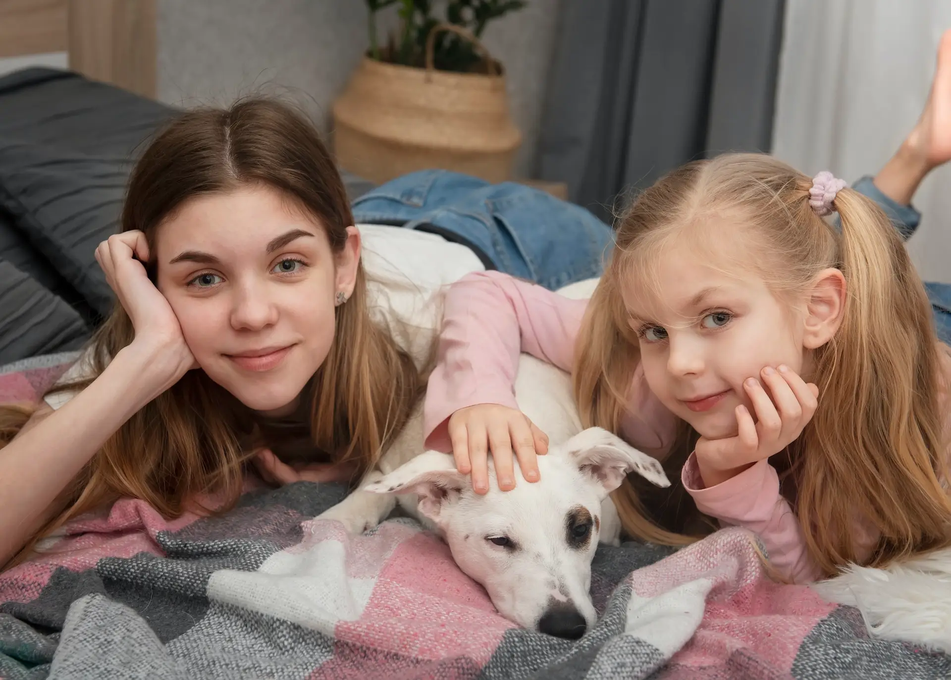 Two young girls lie on a bed, smiling and resting their heads on their hands, while gently petting a white dog lying between them on a cozy blanket. STG-Health