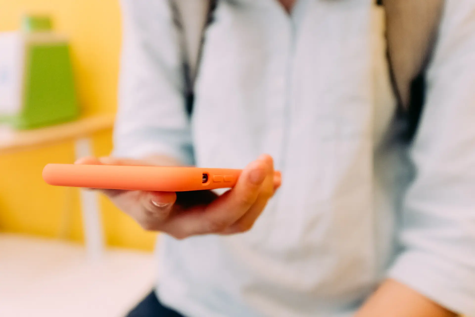 A person wearing a light blue shirt holds an orange smartphone in one hand, with a blurred yellow background and green object in the distance. The persons face is not visible. STG-Health