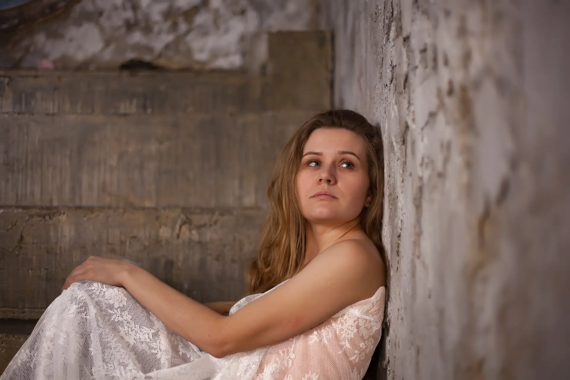 A woman in a white lace dress sits on concrete steps, leaning against a textured, worn wall. She gazes thoughtfully into the distance, with a contemplative and slightly somber expression. STG-Health