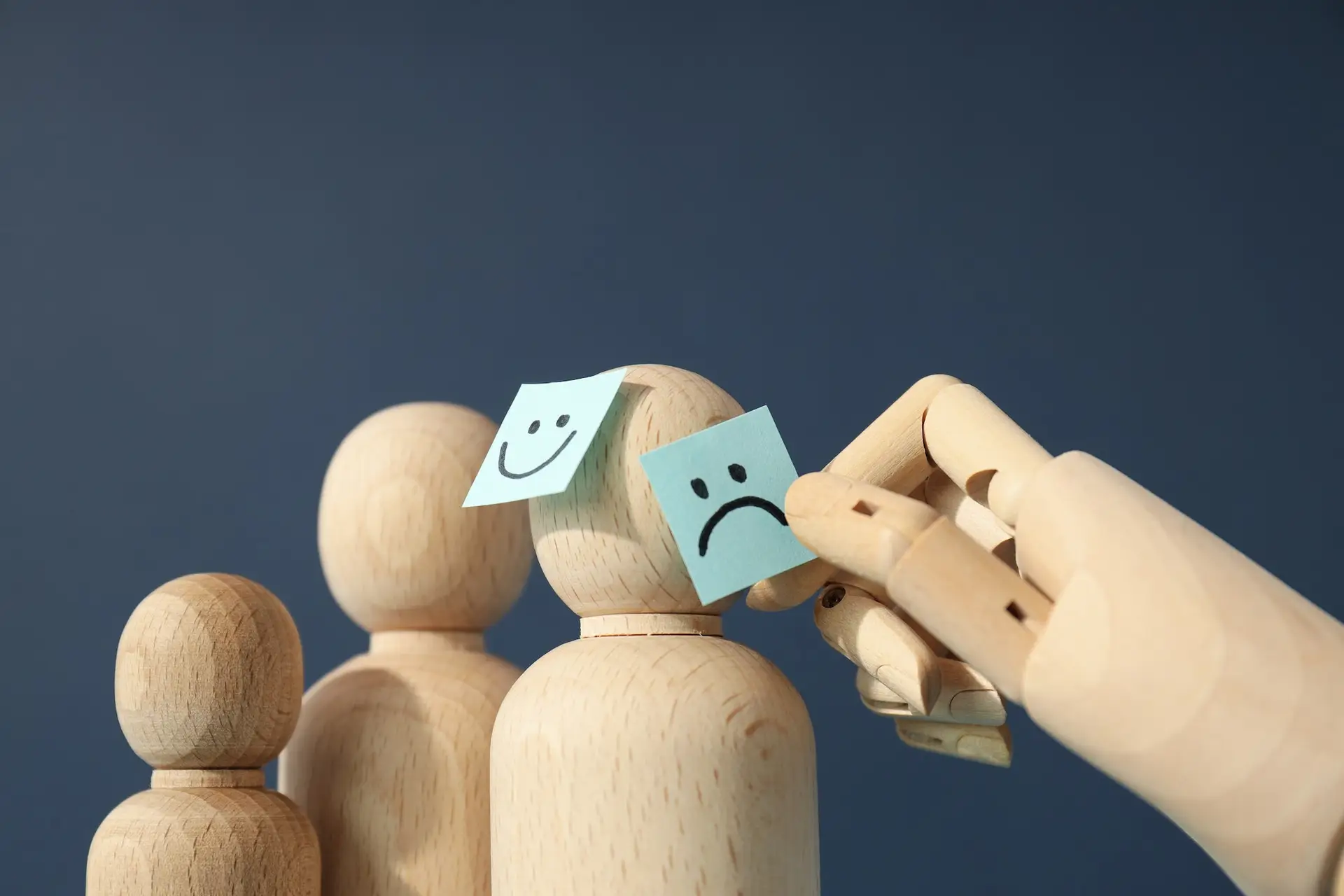 A wooden hand replaces a sad face sticky note with a happy face sticky note on a wooden figure, with more wooden figures standing in the background against a plain dark blue backdrop. STG-Health