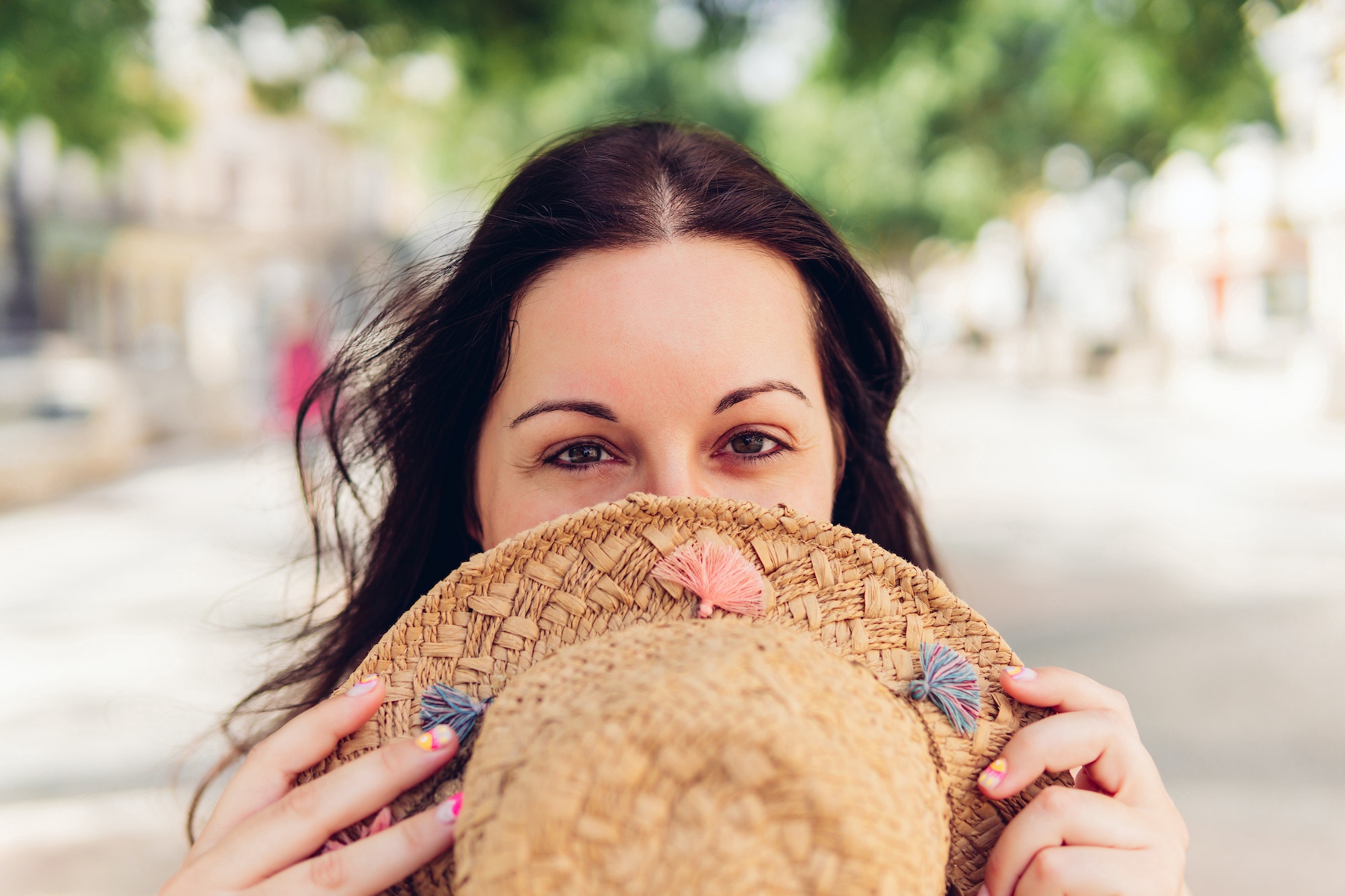 Positive woman hiding face behind hat on street